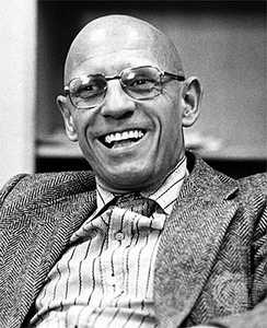 A black and white picture of Michel Foucault. Foucault is a bald man wearing glasses, a blazer, and a striped dress shirt. He is pictured laughing.