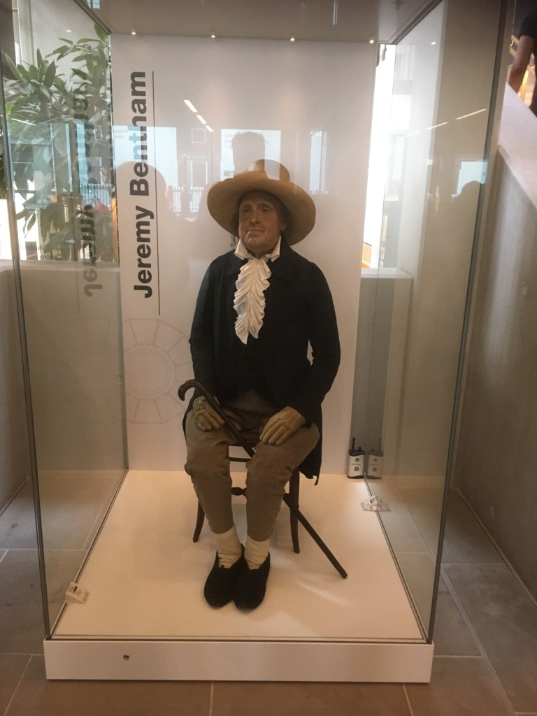 Jeremy Bentham's "Auto-Icon" at The Student Centre in University College London. The Icon appears to be a model of a man wearing old fashioned clothes, including gloves and a hat. He is sitting on a wooden chair with a cane set between his legs. The model is inside a glass case.