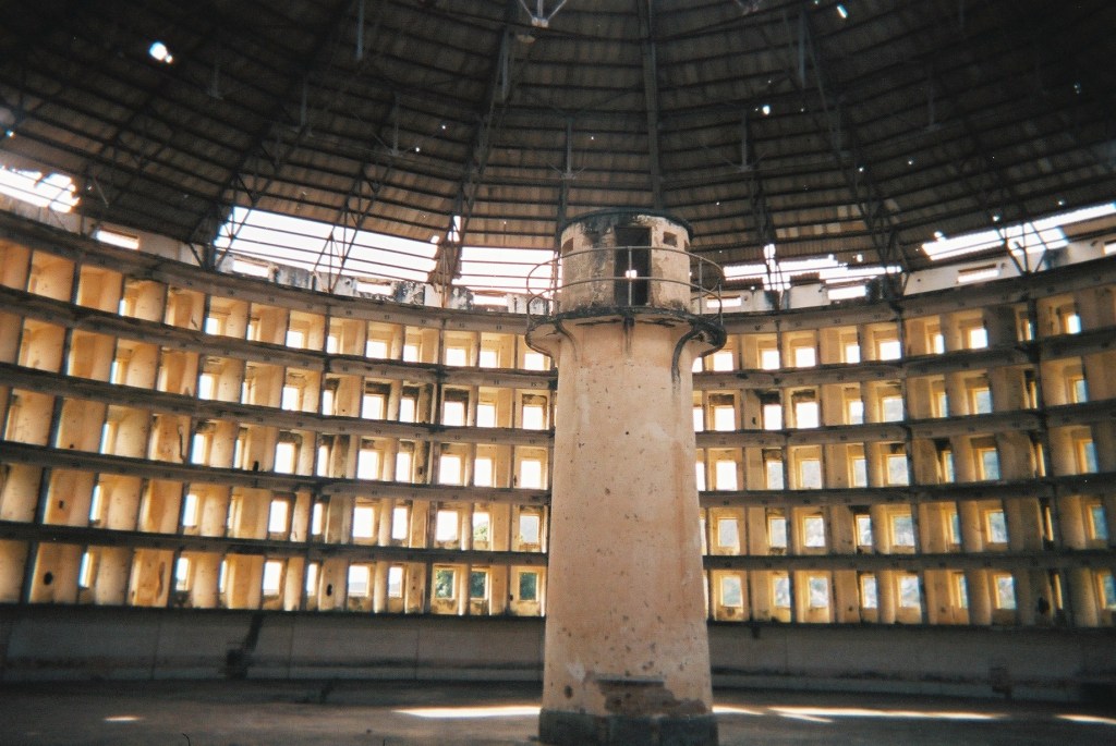 The Presidio Modelo in Cuba. The Presidio is a round building in a state of disrepair. The outer ring appears to consist of rectangular open windows, while a guard tower stands in the center.