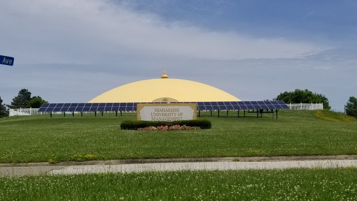 A large, low lying golden dome lies behind a row of solar panels. In the front, a sign reads Maharishi University of Management.