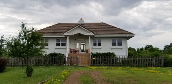 A small white house with a brown roof. It's most notable feature is a small roof ornament above the center of the house. It is also surrounded by a wooden fence.